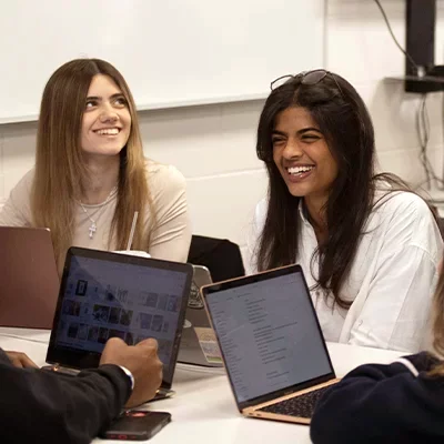 Two female college students sitting at table with laptops and laughing